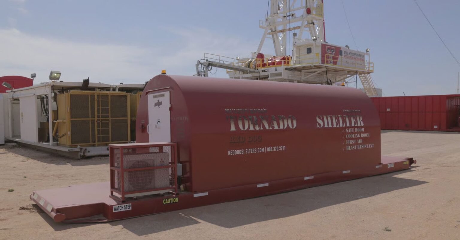 A red, industrial-grade Red Dog tornado shelter sits on a heavy-duty steel skid at a dusty outdoor job site.